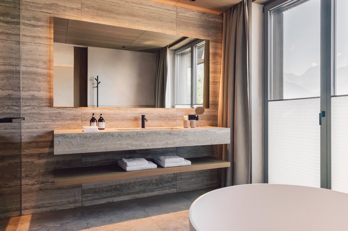 Badezimmer im Luxury Penthouse 2 in Fiss Ein modernes Badezimmer mit Waschtisch, großer Spiegelwand und freistehender Badewanne am Fenster.