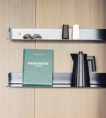 Regal mit Designobjekten im Apartment Ein Regal aus hellem Holz mit Buch, Kanne, Bechern und Sonnenbrille.
