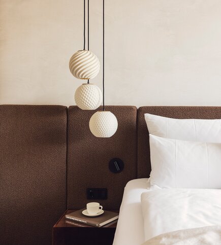 Schlafzimmer mit Design-Pendelleuchten Ein Bett mit weißen Kissen steht neben einem Nachttisch mit Tasse und Buch unter drei Design-Pendelleuchten.